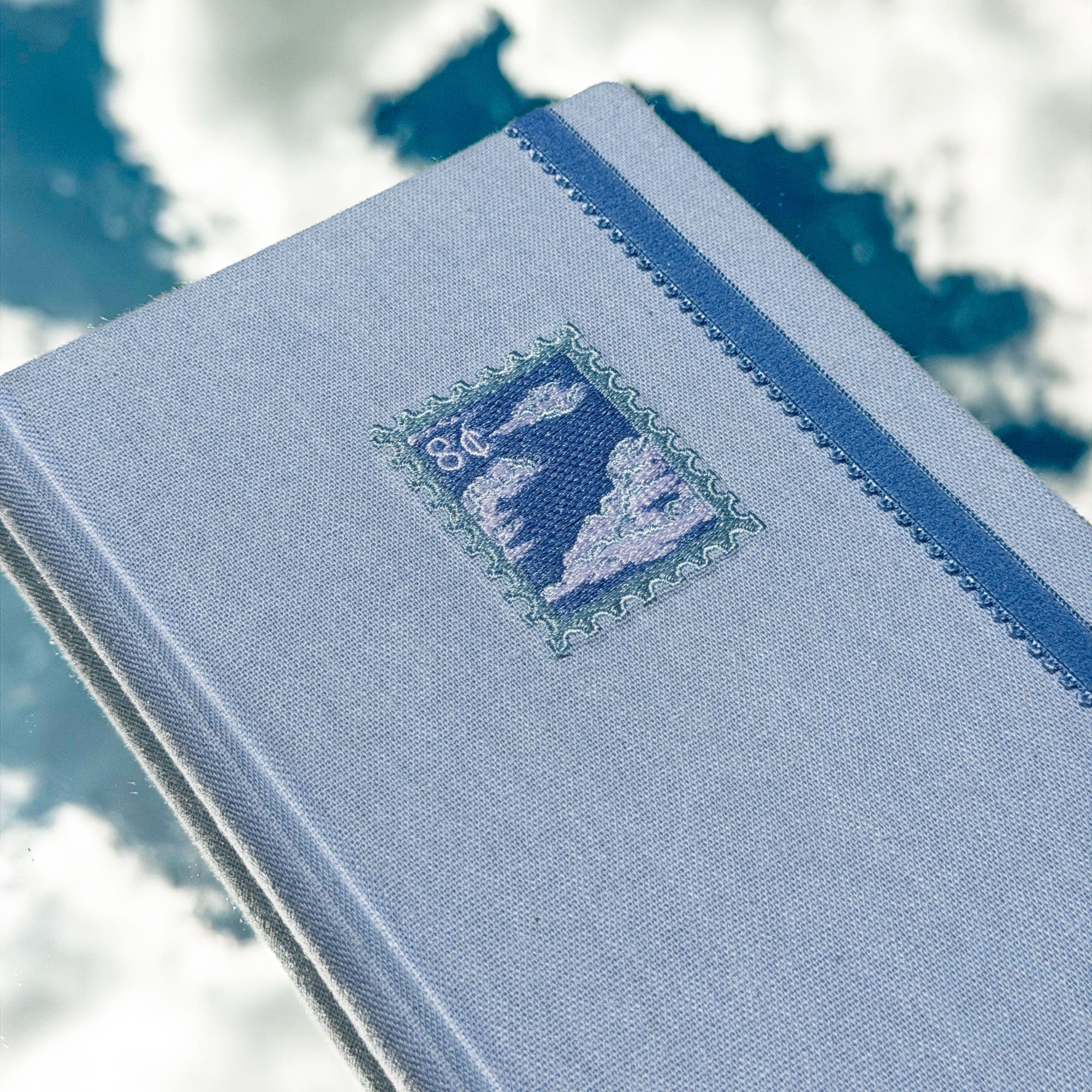 Primer plano de la portada del journal Trazos de cielo: la estampilla de nubes bordada sobre la tela azul, con la cinta elástica al costado.